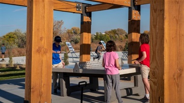 A group of kids play ping pong on a concrete table underneath a covering