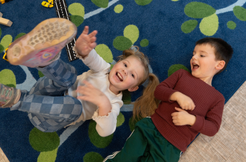 Two kids smile while playing on a carpet in a classroom