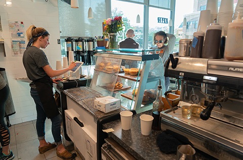 A person taking an order at a coffee shop with customers in the store and an espresso machine in the foreground