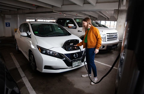 A woman charges a white electric vehicle (Nissan Leaf) at a public EV charging station inside a parking garage in Fort Collins, Colorado.