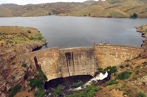 Aerial view of Halligan Dam and Reservoir, looking northwest. The water is gray and the dam is brown. Hills are in the background.